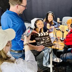 Drei Personen sitzen in einem Tonstudio mit Kopfhörern vor Mikrofonen und lächeln. Eine Person hält eine Filmklappe und ein Handy mit laufender Kamera. Im Hintergrund steht eine Person mit blauer Jacke und eine weitere Person mit roter Jacke.