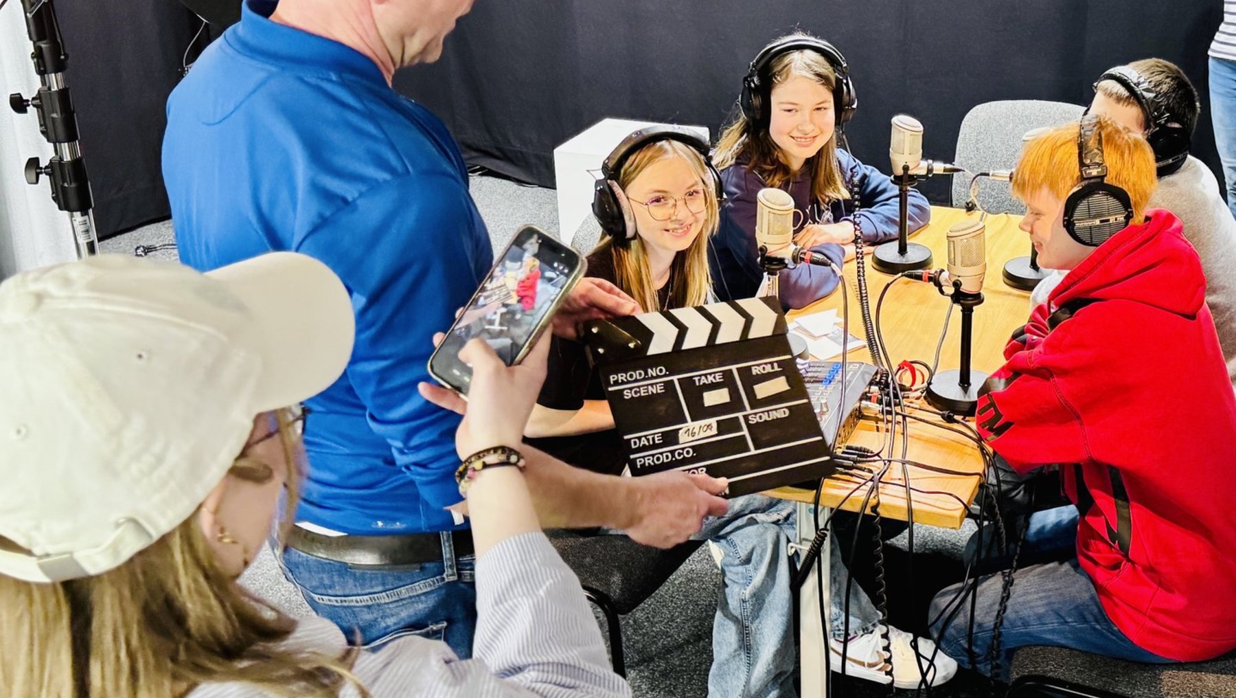 Drei Personen sitzen in einem Tonstudio mit Kopfhörern vor Mikrofonen und lächeln. Eine Person hält eine Filmklappe und ein Handy mit laufender Kamera. Im Hintergrund steht eine Person mit blauer Jacke und eine weitere Person mit roter Jacke.