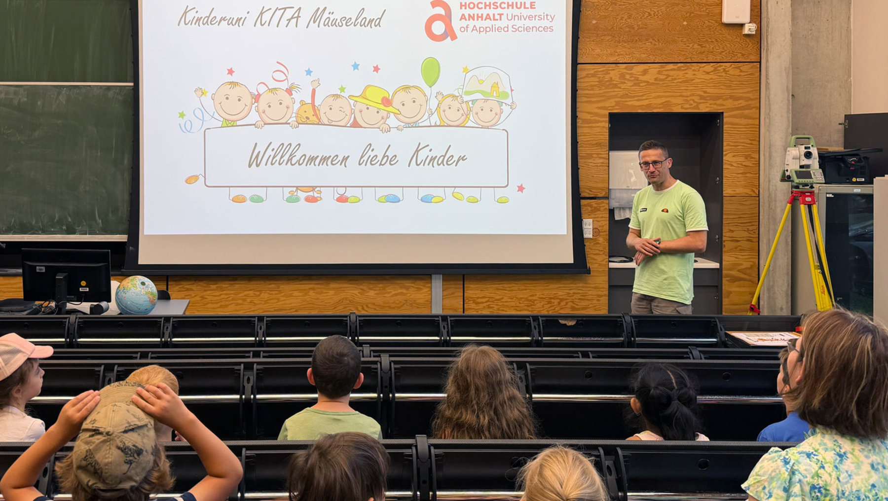 Ein Mann steht vor einem Publikum aus Kindern in einem Hörsaal und hält eine Begrüßungsrede. Hinter ihm ist eine Präsentation mit dem Text „Willkommen liebe Kinder“ und kindlichen Illustrationen zu sehen.