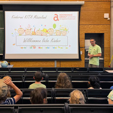 Ein Mann steht vor einem Publikum aus Kindern in einem Hörsaal und hält eine Begrüßungsrede. Hinter ihm ist eine Präsentation mit dem Text „Willkommen liebe Kinder“ und kindlichen Illustrationen zu sehen.