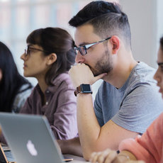 Eine Gruppe von Studierenden sitzt in einem Raum und arbeitet konzentriert an Laptops. Vier Personen sind zu sehen, zwei Frauen und zwei Männer. Sie scheinen in einem Seminar oder einer Vorlesung zu sein und sind auf ihre Bildschirme fokussiert.