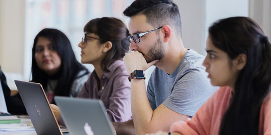 Eine Gruppe von Studierenden sitzt in einem Raum und arbeitet konzentriert an Laptops. Vier Personen sind zu sehen, zwei Frauen und zwei Männer. Sie scheinen in einem Seminar oder einer Vorlesung zu sein und sind auf ihre Bildschirme fokussiert.