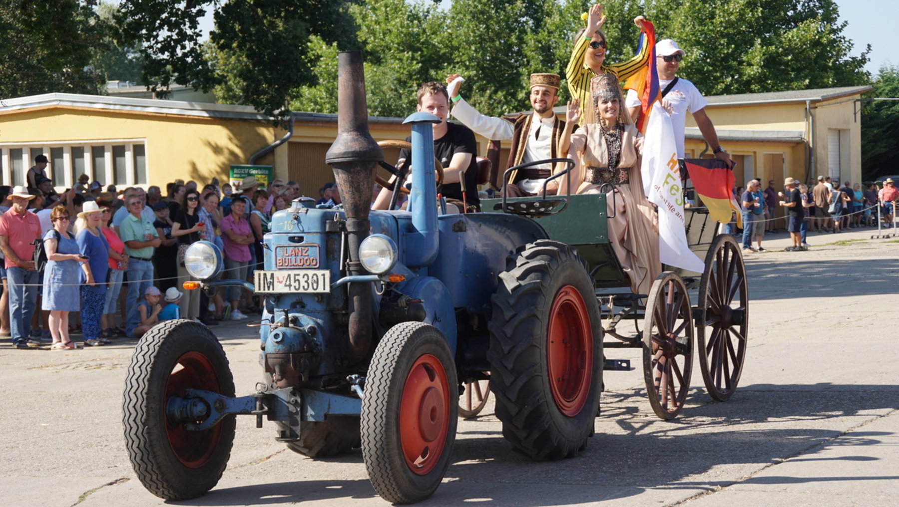 Vier Personen fahren auf einem alten blauen Traktor mit rot lackierten Reifen und angehängtem zweirädrigem Wagen durch eine Straße. Sie winken und halten eine deutsche Flagge sowie ein buntes Tuch. Im Hintergrund steht ein Publikum hinter Absperrungen.