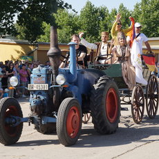Vier Personen fahren auf einem alten blauen Traktor mit rot lackierten Reifen und angehängtem zweirädrigem Wagen durch eine Straße. Sie winken und halten eine deutsche Flagge sowie ein buntes Tuch. Im Hintergrund steht ein Publikum hinter Absperrungen.