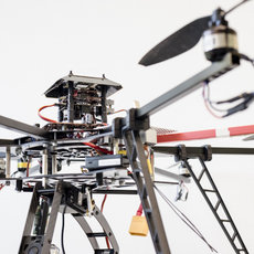 A close-up of a multi-rotor drone with four propellers, showing its central frame, wiring, and mounted sensors or camera equipment. The drone has a sturdy landing gear structure.