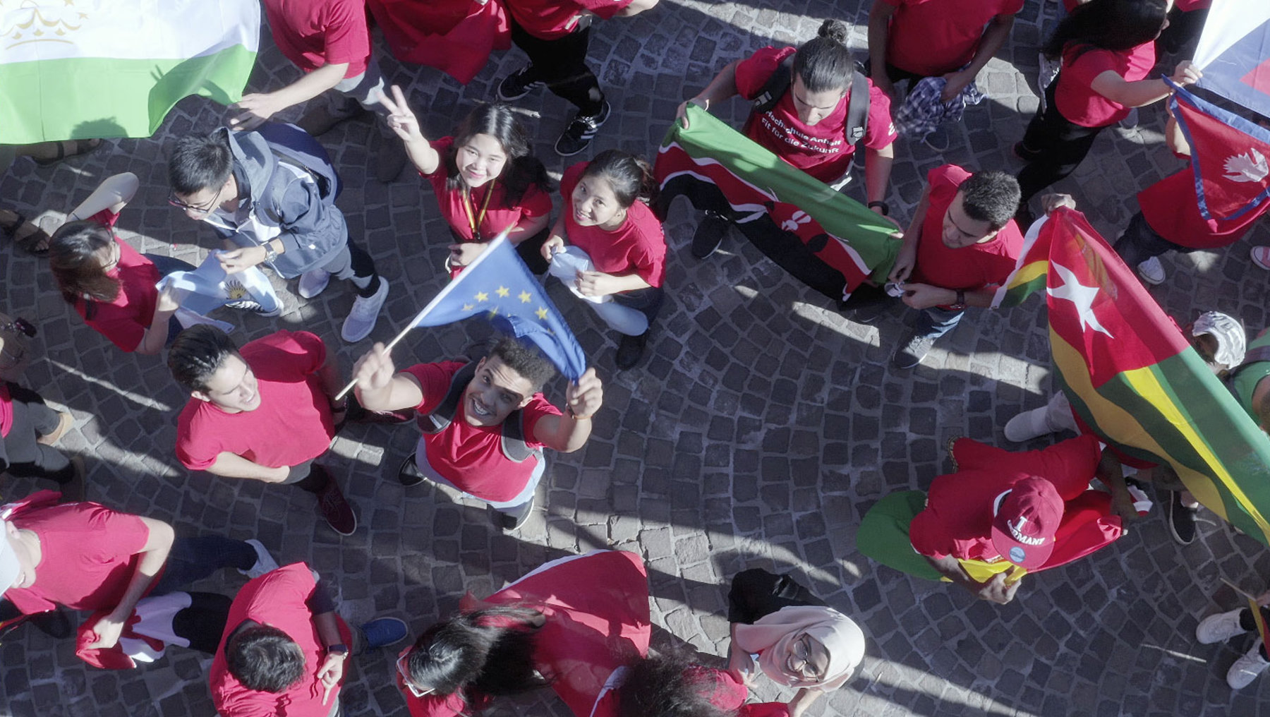Eine Gruppe von Kindern in roten T-Shirts sitzt auf dem Boden und hält Flaggen. Sie scheinen an einer Veranstaltung oder einem Fest teilzunehmen. Die Flaggen haben verschiedene Farben und Muster. Die Kinder wirken fröhlich und engagiert.