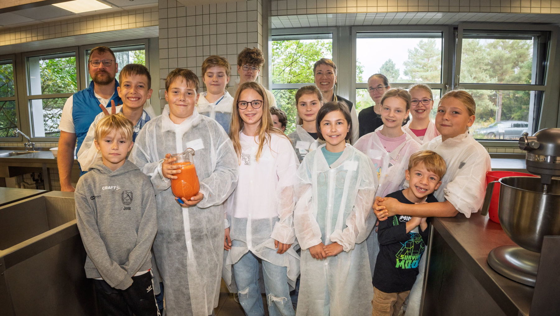 Gruppe von Kindern und Erwachsenen in einer Küche, alle in weißen Schutzanzügen und Hauben. Die Kinder halten verschiedene Kütengeräte, alle lächeln fröhlich für ein Gruppenfoto.