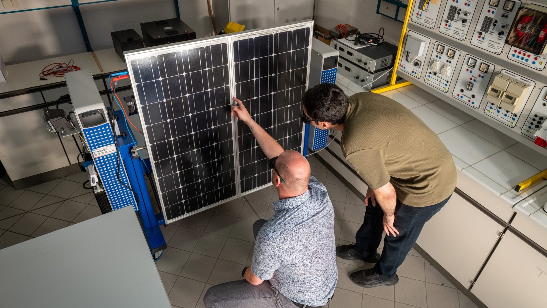 Zwei Männer in einem Labor der Hochschule Anhalt zur Photovoltaik-Forschung: einer kniet und zeigt auf ein großes, senkrecht stehendes Solarmodul, während der andere danebensteht und auf die Zellen deutet. Im Hintergrund technische Geräte und Messinstrumente.
