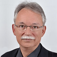 A man with short gray hair, glasses, and a mustache. He is wearing a dark vest over a collared shirt and is looking directly at the camera with a neutral expression. The background is plain and white.