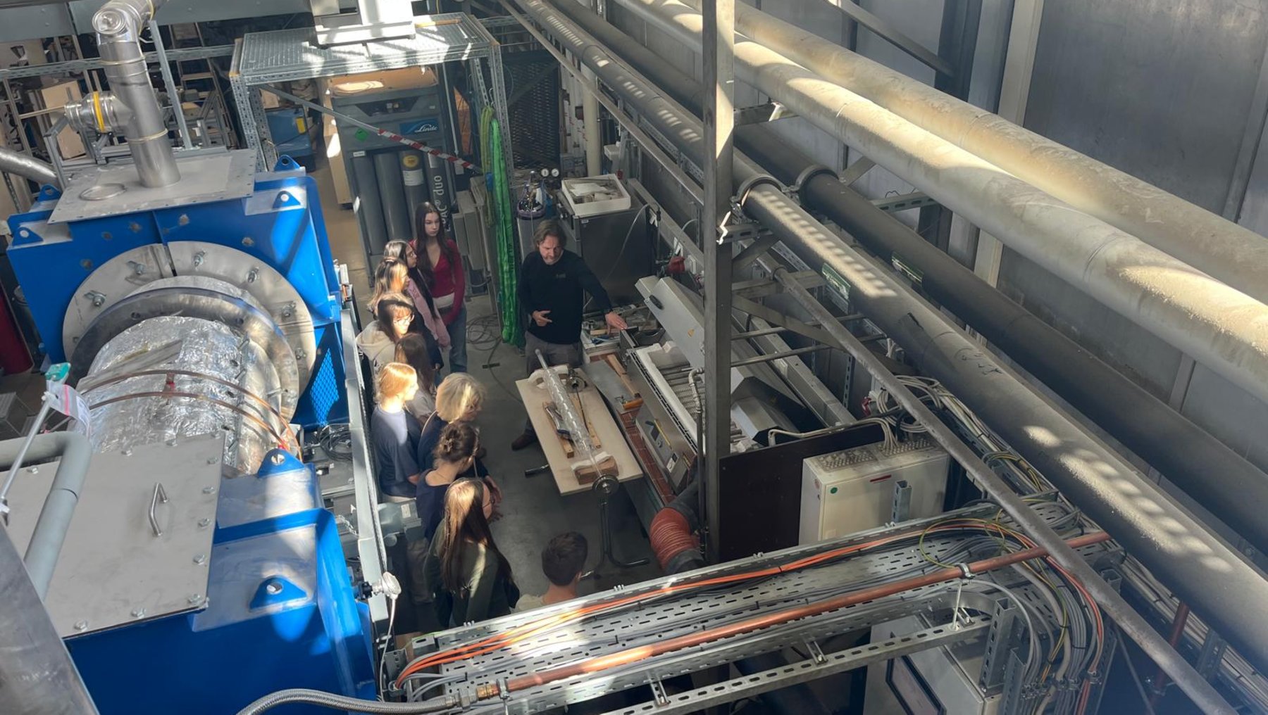 Großer technischer Raum mit mehreren Personen, die eine komplexe Maschinenanlage mit Rohrleitungen, Ventilen und blauen Komponenten betrachten. Im Vordergrund eine große, runde Apparatur, im Hintergrund ein langer, metallischer Aufbau mit farbigen Kabeln und Messinstrumenten.