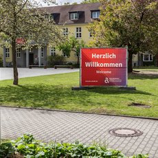 "Ein Campusgelände mit einem roten Schild, das 'Herzlich Willkommen' auf Deutsch und 'Welcome' auf Englisch schreibt. Im Hintergrund befinden sich Gebäude und Bäume. Ein weißes Schild mit der Nummer 08 und einem Text steht links im Bild."