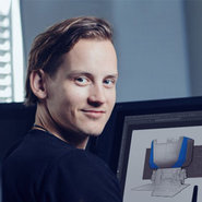 Ein lächelnder Mann mit kurzem braunem Haar sitzt vor einem Bildschirm, auf dem eine 3D-Darstellung eines Gebäudes oder einer Architekturstruktur zu sehen ist.