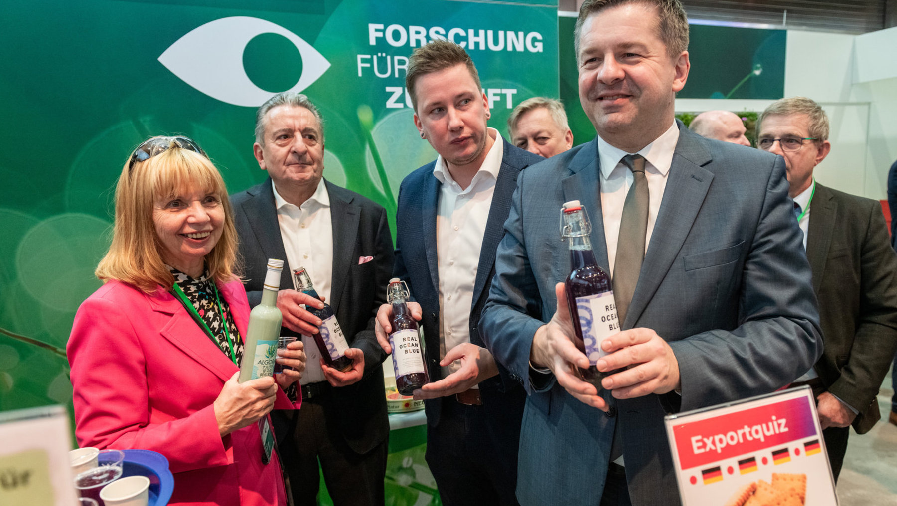 Vier Personen auf einer Messe stehend, die jeweils eine Flasche Bier der Marke „Real Ocean Blue“ halten. Im Hintergrund steht ein Stand mit dem Schriftzug „Forschung für Zukunft“ und einem großen Auge-Logo.