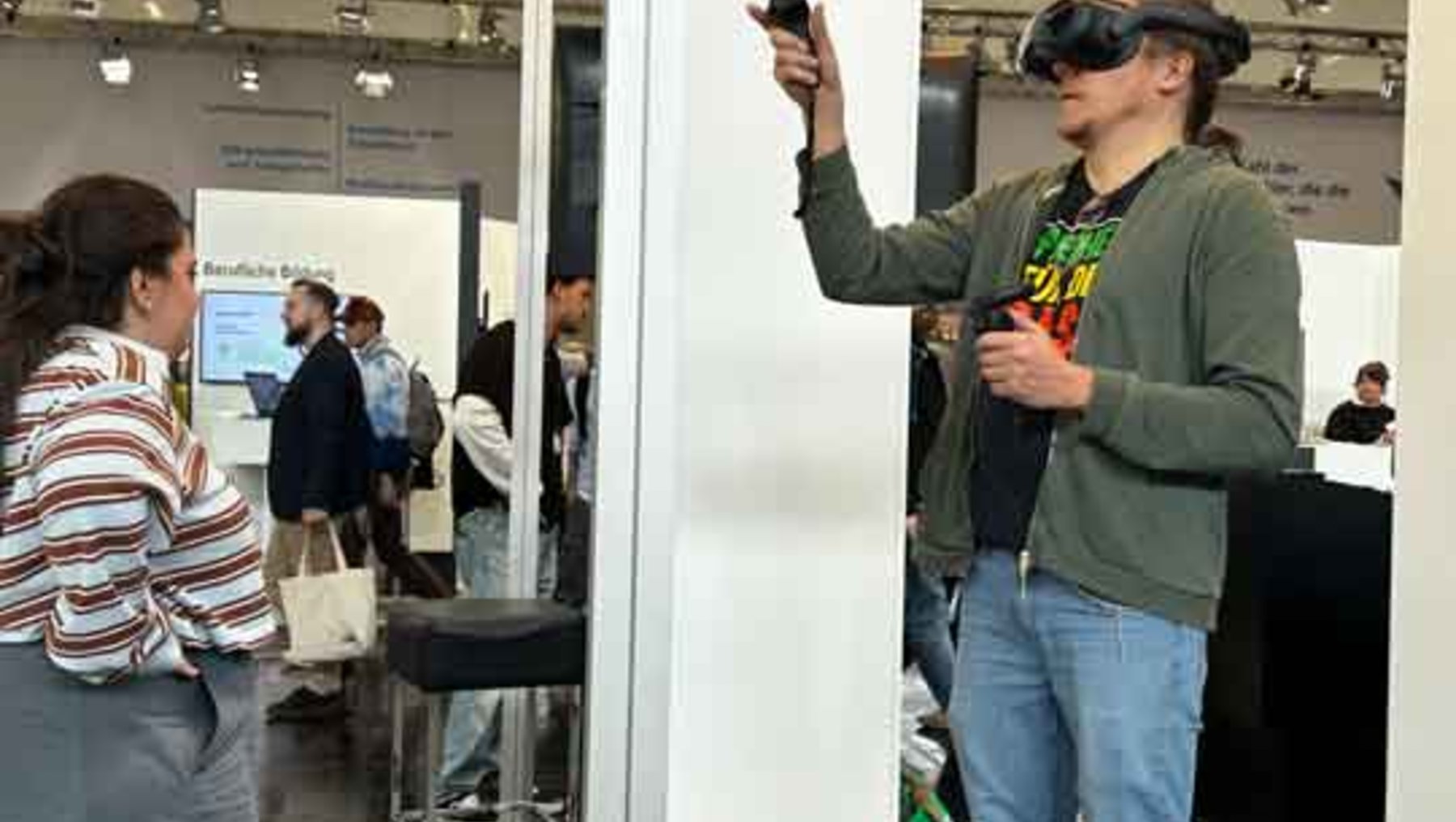 Ein Mann in grünem Pullover und Jeans hält ein Smartphone in der Hand und nutzt gleichzeitig eine Virtual-Reality-Brille, während er auf eine weiße Säule oder ein Display zeigt. Im Hintergrund sind weitere Messebesucher zu sehen.