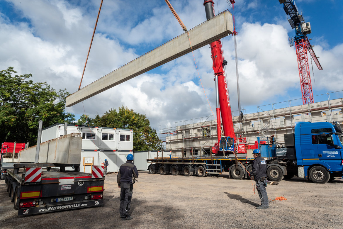 Zwei Bauarbeiter in Schutzausrüstung überwachen das Einbauen einer großen Betonstütze mit einem roten Kransystem auf einer Baustelle. Im Hintergrund sind ein Gerüst, ein weißer Container und ein blauer Lkw mit Anhänger zu sehen.