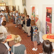 Gruppe älterer Menschen in einem hellen Raum mit Ausstellungswänden, die sich unterhalten und an kleinen Tischen mit Gläsern und Flaschen stehen. Einige betrachten Plakate und Bilder an den Wänden.