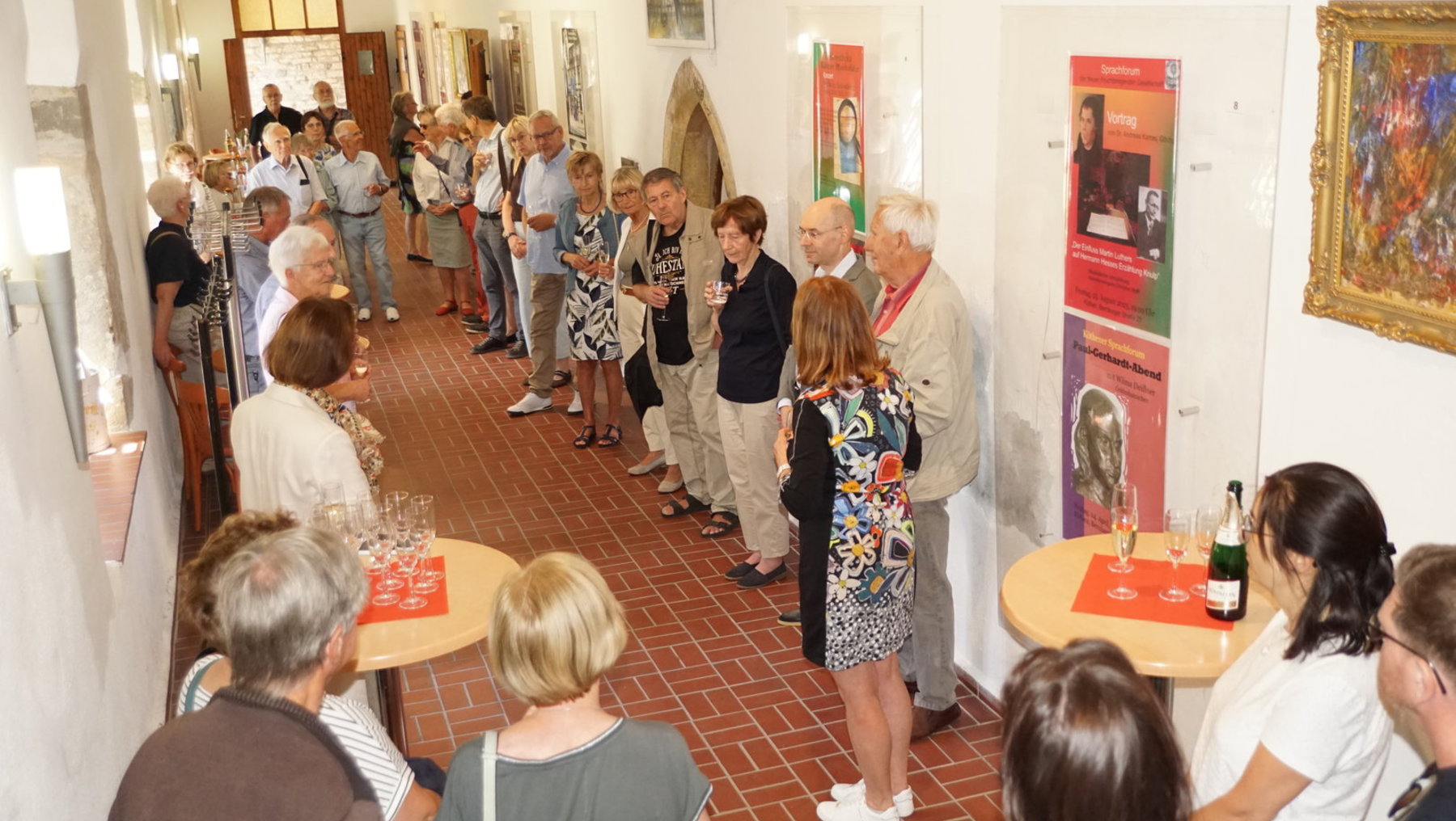 Gruppe älterer Menschen in einem hellen Raum mit Ausstellungswänden, die sich unterhalten und an kleinen Tischen mit Gläsern und Flaschen stehen. Einige betrachten Plakate und Bilder an den Wänden.