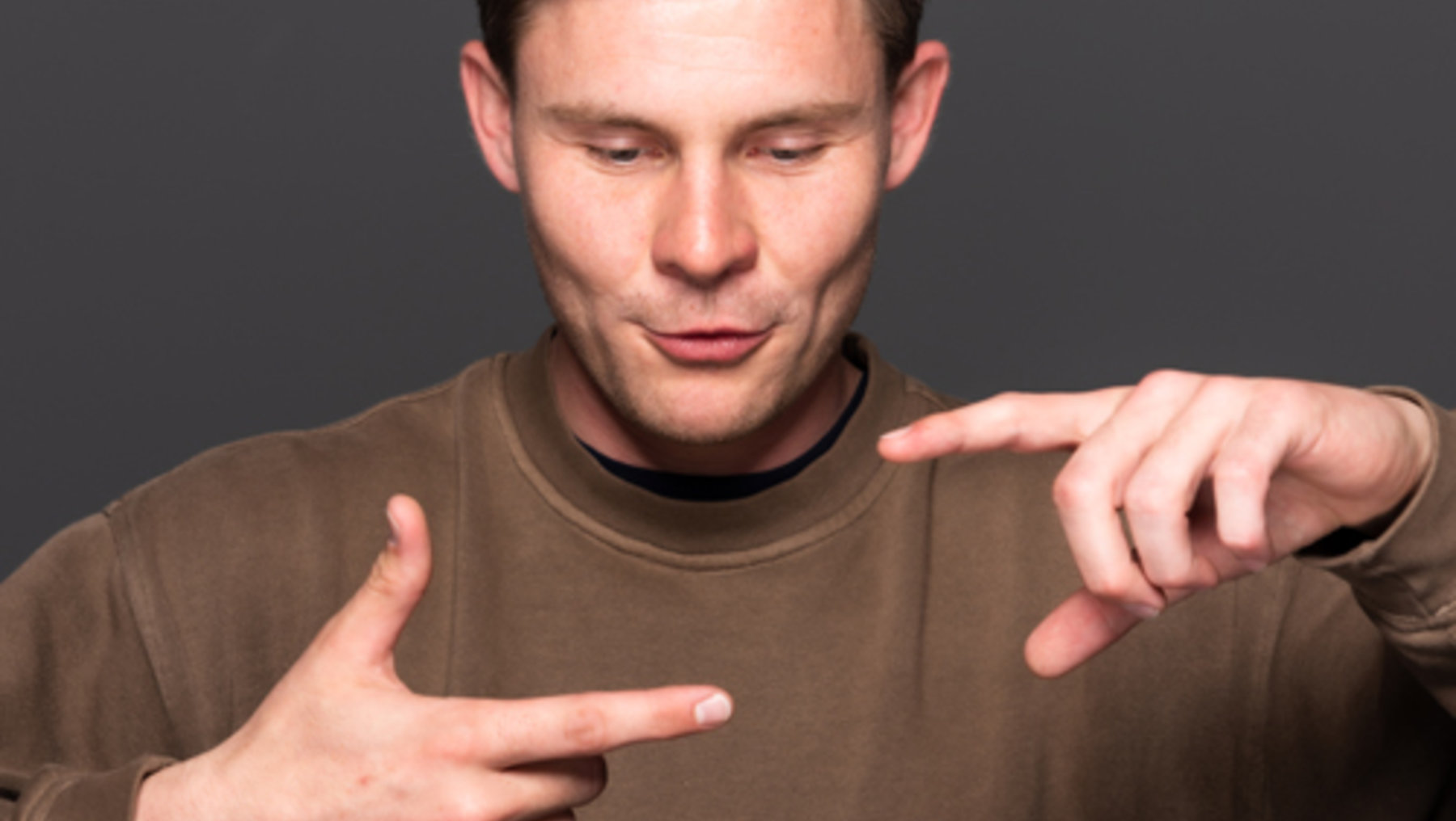 Ein Mann in einem braunen Adidas-Pullover zeigt mit beiden Händen nach außen, als würde er etwas präsentieren oder eine Geste machen. Er blickt konzentriert nach unten.
