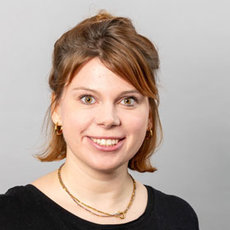 Ein lächelndes Porträt einer Person mit kurzen roten Haaren, die zu einem Dutt gebunden sind. Die Person trägt ein schwarzes Rollkragen-Shirt und goldene Ohrringe sowie eine Kettenhalskette. Das Bild ist neutral und professionell.