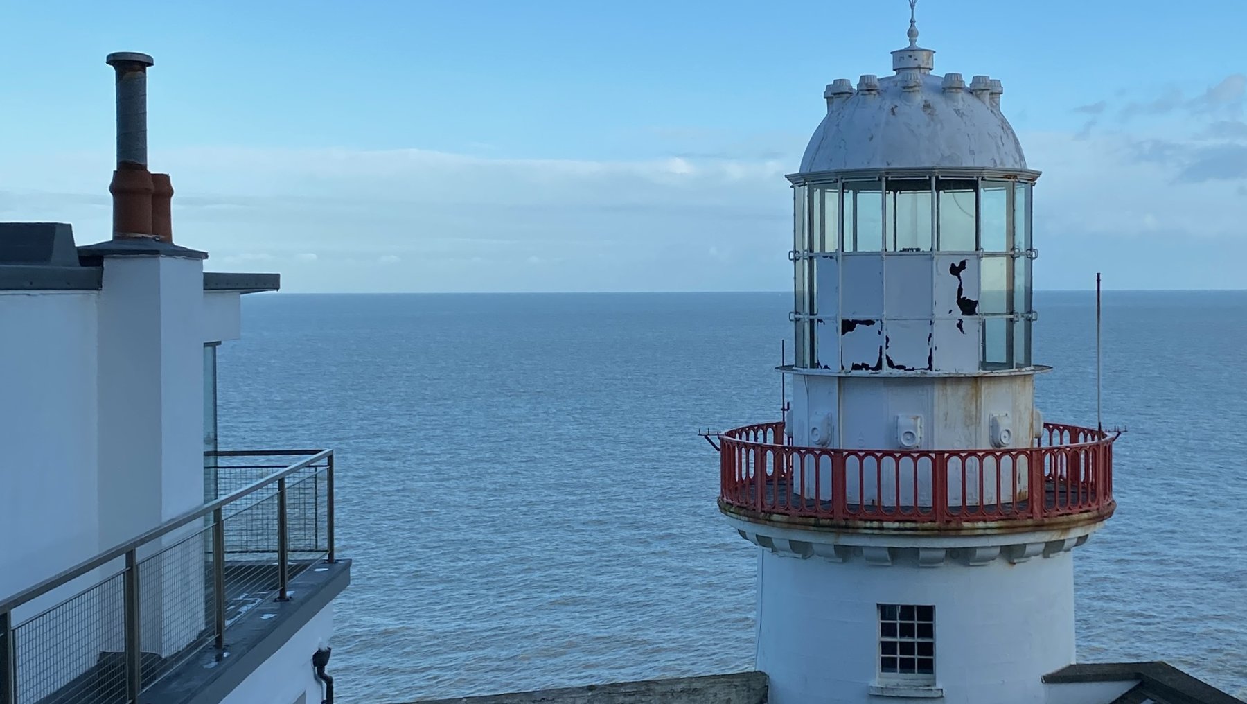 Leuchtturm mit weißem Turm und roter Galerie, der auf einer steinigen Klippe am Meer steht. Im Hintergrund ist das offene Wasser mit leicht bewölktem Himmel zu sehen. Ein Treppenaufgang führt zur Plattform mit Geländer.