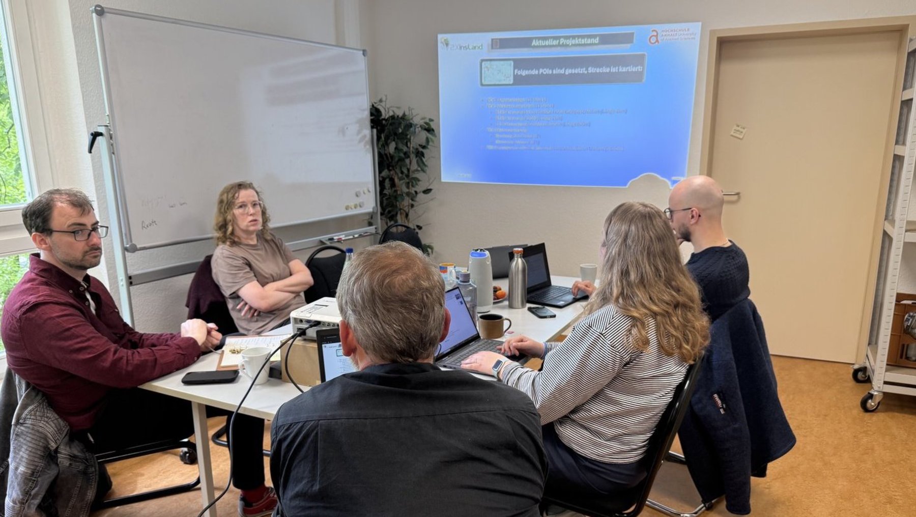 Fünf Personen sitzen in einem Besprechungsraum an einem Tisch, arbeiten an Laptops und diskutieren. Im Hintergrund ist eine Leinwand mit einem Präsentationsthema zu sehen, das mit „digitale Transformation“ beginnt.