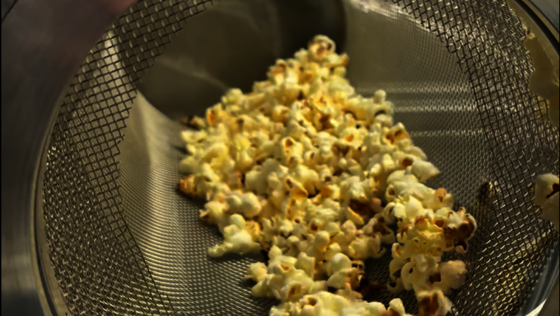 Frischer, knuspriger Popcorn-Haufen in einem Metall-Sieb, leicht goldbraun und ungleichmäßig geformt, mit einigen kleinen Klümpchen.