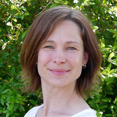 A woman with shoulder-length brown hair is smiling gently. She is standing in front of a lush green background, possibly foliage or a garden. She is wearing a light-colored top. The image captures a serene and pleasant moment.