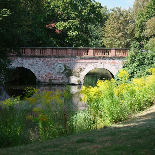 An old stone bridge with two arches spans over a small body of water. The bridge is surrounded by lush greenery and trees, creating a serene and natural setting. The water reflects the bridge and the surrounding vegetation.