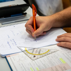Eine Person hält einen roten Stift in der Hand und schreibt in ein kariertes Heft, während ein Geodreieck daneben liegt. Im Hintergrund sind Notizen und ein Taschenrechner zu sehen.