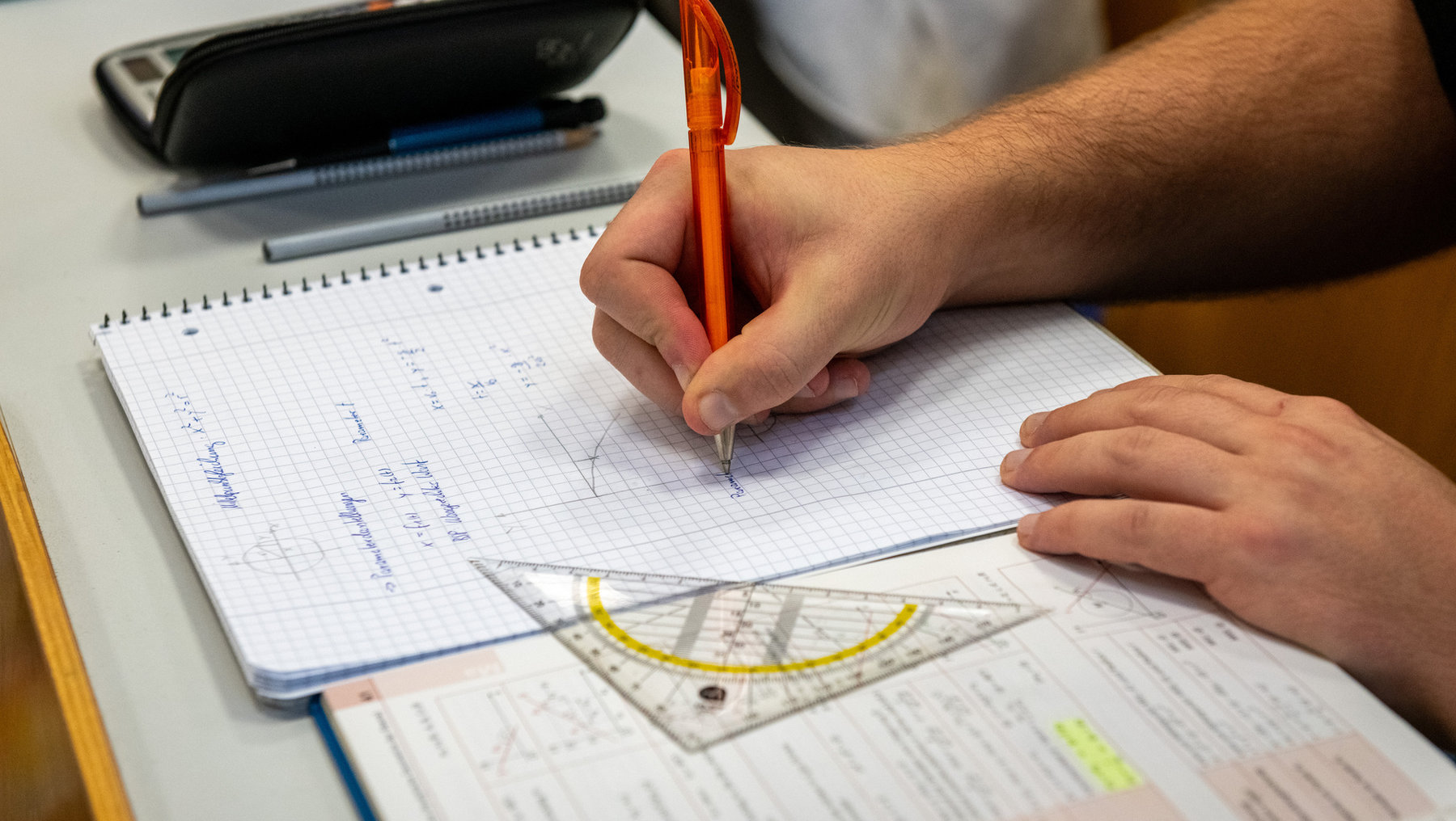 Eine Person hält einen roten Stift in der Hand und schreibt in ein kariertes Heft, während ein Geodreieck daneben liegt. Im Hintergrund sind Notizen und ein Taschenrechner zu sehen.