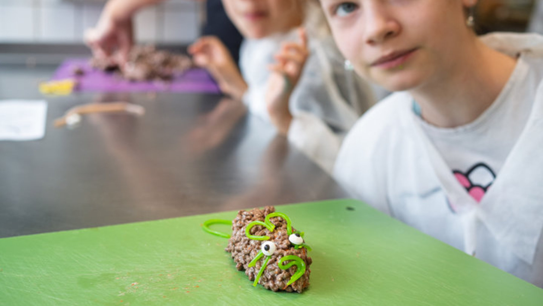Zwei Kinder basteln gemeinsam an einer Tischplatte. Im Vordergrund liegt ein grünes Plakat mit einer selbstgebastelten Figur aus buntem Papier, die wie ein Tier mit großen Augen aussieht. Die Kinder wirken konzentriert und zufrieden.