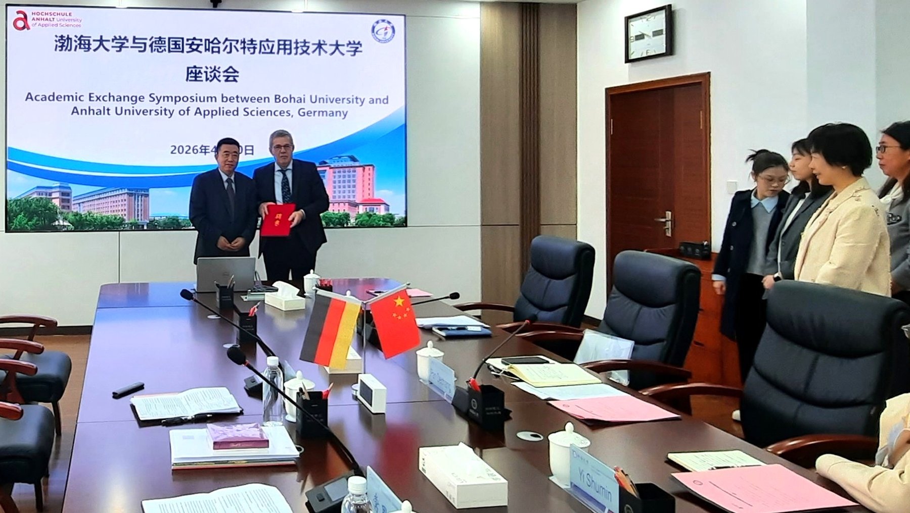 Zwei Männer stehen an einem Konferenztisch und halten jeweils eine Flagge – eine deutsche und eine chinesische – während sie ein rotes Dokument überreichen. Im Hintergrund sitzen weitere Personen in einem Seminarraum, der für ein akademisches Austauschtreffen zwischen Bohai-Universität und der Anhalt-Universität vorbereitet ist.