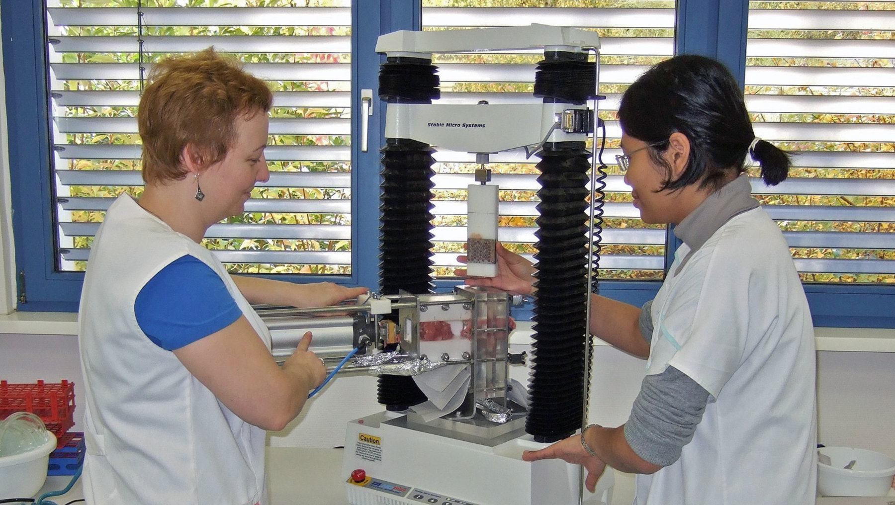 Zwei Personen untersuchen gemeinsam eine Prüfmaschine mit koaxialer Anordnung, bei der wahrscheinlich Materialien auf Haltbarkeit oder Belastungsfestigkeit getestet werden. Beide scheinen fokussiert mit den Gerätekomponenten interagieren.')