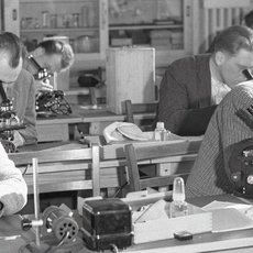 A black-and-white image of several people in a laboratory setting, intently working with microscopes and other scientific equipment. They appear to be engaged in research or study, with papers and tools on their desks. The room is filled with individuals focused on their tasks.