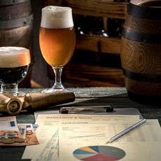 Die Abbildung zeigt zwei Gläser mit Bier, eines dunkel und eines hell. Daneben liegen Hopfenzapfen, Bierrezepte mit Etiketten und ein Blatt mitehiclem Grafiken sowie Zahlen, möglicherweise eine Analyse oder Kostenaufstellung zum Bierbrauen. Ein Stift Completed den Szene auf einem Tisch.