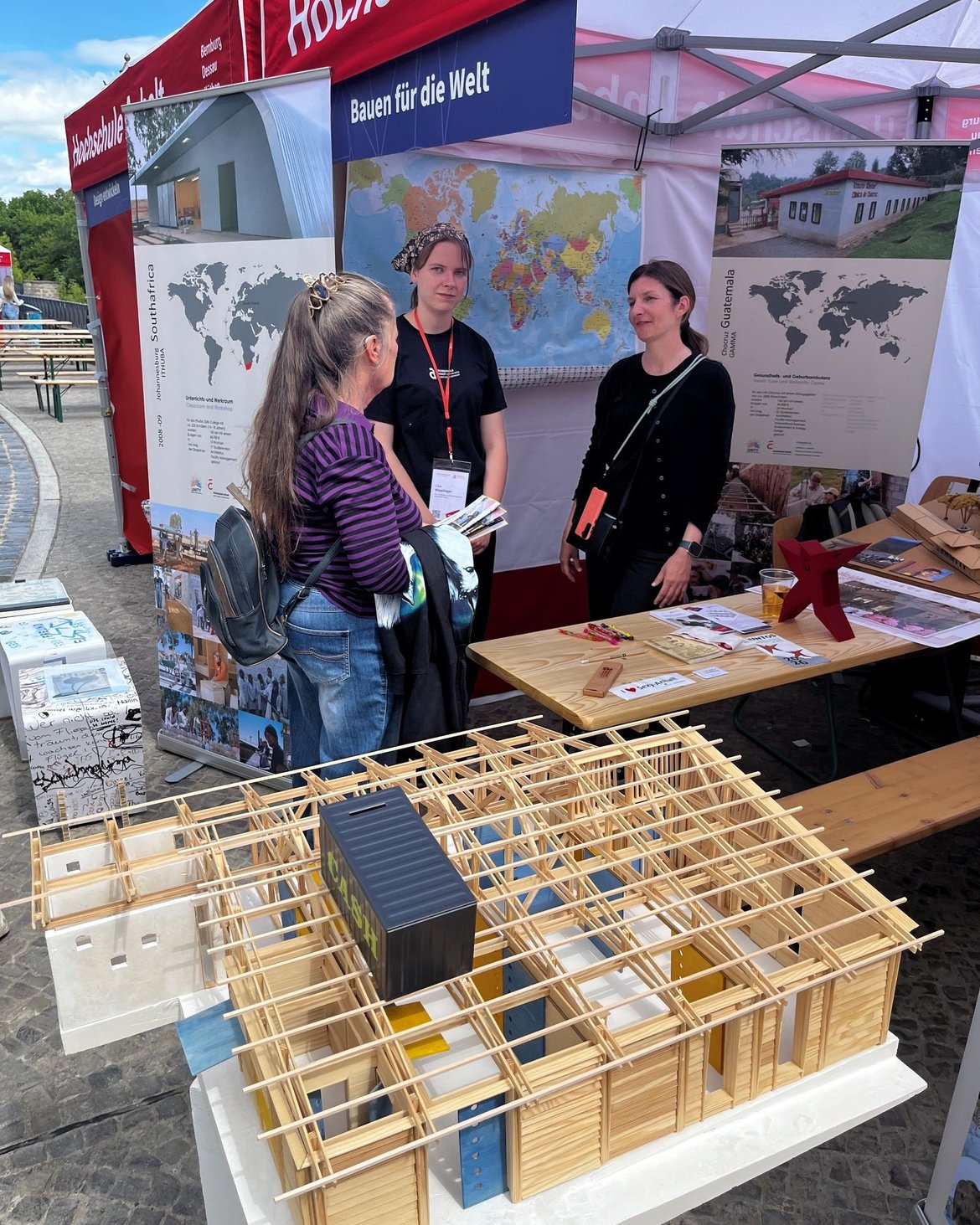 Drei Personen stehen an einem Infostand mit dem Thema „Bauen für die Welt“. Vor ihnen liegt ein großes Holzmodell eines Gebäudes mit verschiedenen farbigen Elementen, das nachhaltiges Bauen oder eine Bauweise für Entwicklungsprojekte darstellt.