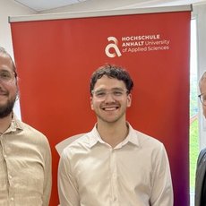 Drei Männer stehen lächelnd nebeneinander vor einem Banner der Hochschule Ansbach, das das Logo der Hochschule und den Schriftzug „University of Applied Sciences“ zeigt. Der mittlere Mann hält ein Dokument.