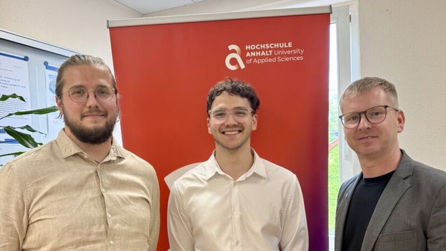 Drei Männer stehen lächelnd nebeneinander vor einem Banner der Hochschule Ansbach, das das Logo der Hochschule und den Schriftzug „University of Applied Sciences“ zeigt. Der mittlere Mann hält ein Dokument.