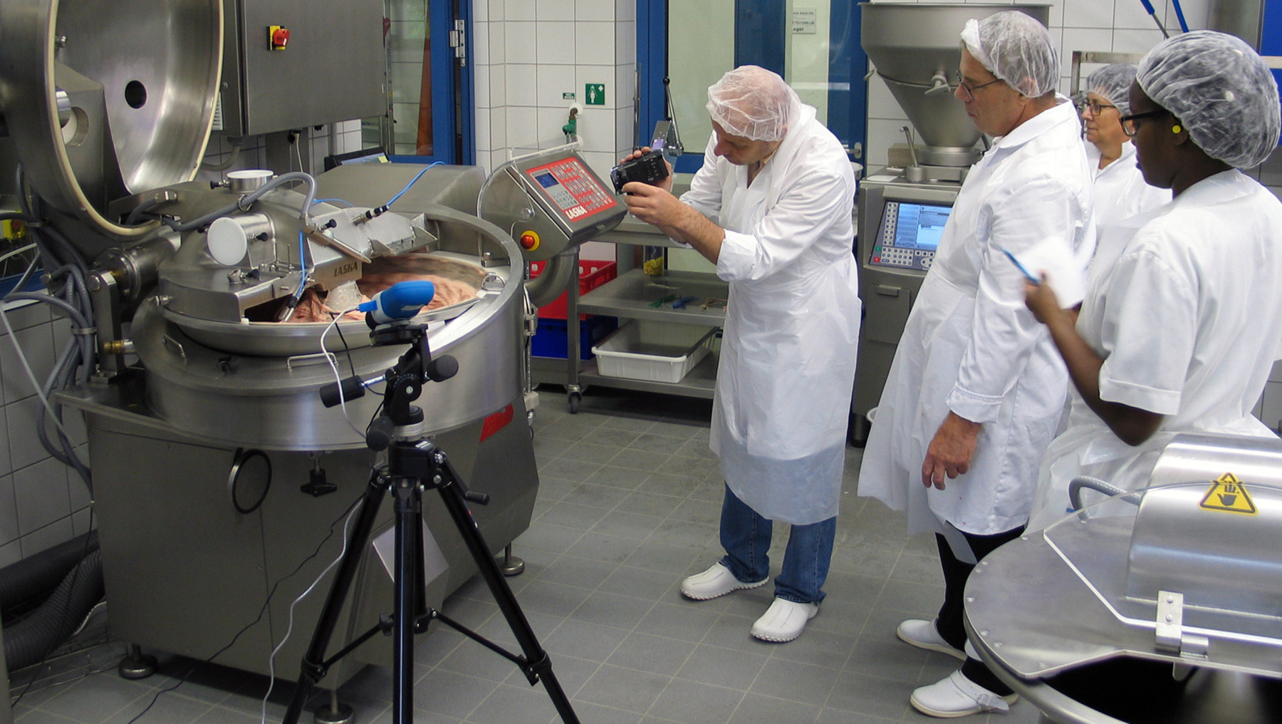 Diese Abbildung zeigt eine Person in weißer Schutzkleidung, die ein industrielles Gerät bedient, während andere im Hintergrund zuschauen. Äußeres Umfeld ist eine Produktionsküche oder ein Reinraum mit Hochtechnikmaschinen. Thematisch geht es um die Demonstration eines Hygieneprozesses.