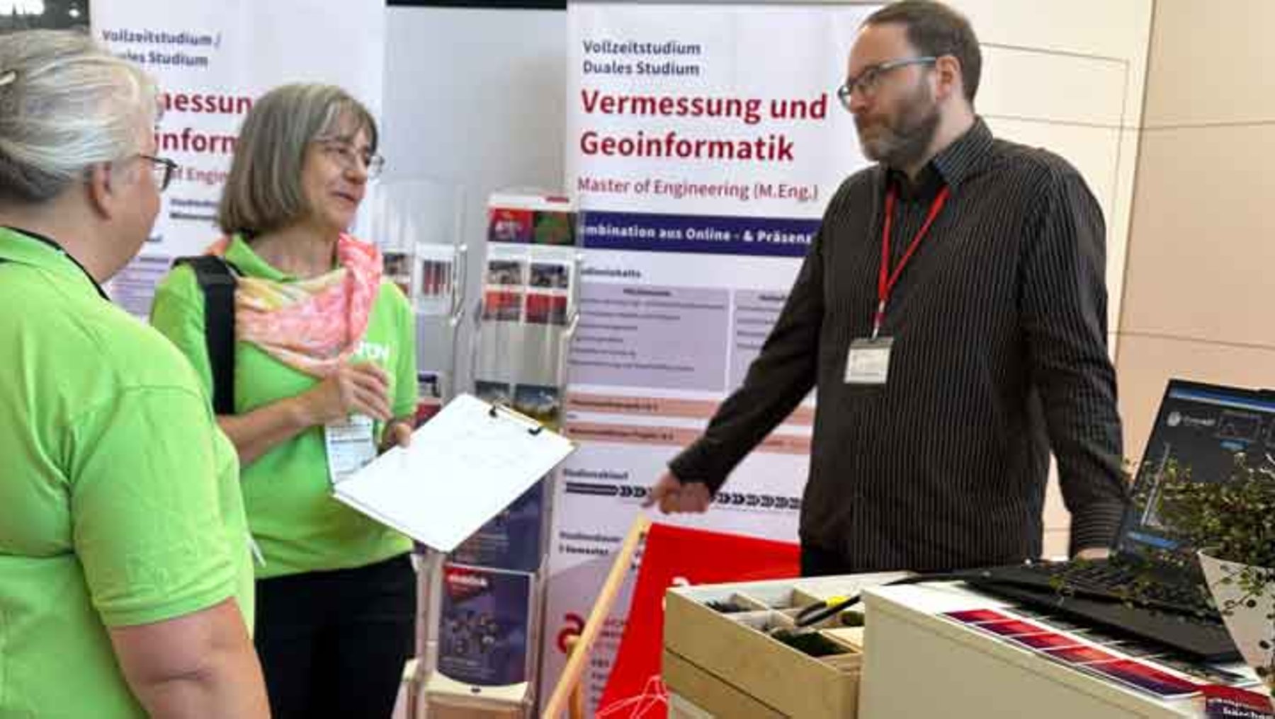 Drei Personen an einem Infostand zu einem Fernstudiengang Vermessung und Geoinformatik an der Dualen Hochschule. Eine Frau hält ein Informationsblatt, ein Mann mit Namensschild erklärt etwas und zeigt auf eine offene Kiste mit Materialien. Im Hintergrund Plakate mit Studieninformationen.