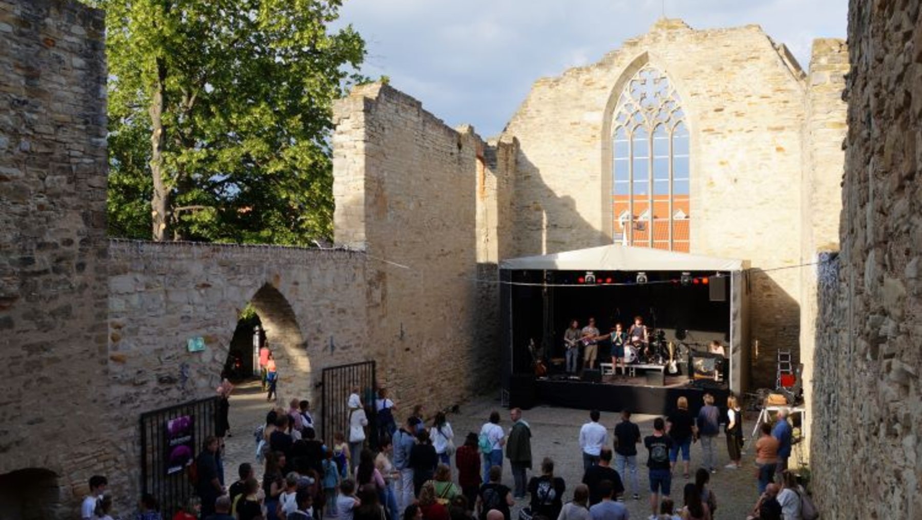 "Eine Menschenmenge versammelt sich in den Ruinen einer alten Kirche mit einer großen Fensterrose. Auf einer Bühne im Hintergrund spielen Musiker. Die Atmosphäre wirkt festlich und lebhaft."