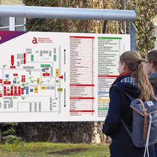Two people are standing outdoors, looking at a large map of "Campus Strenzfeld." The map shows buildings, pathways, and various facilities within the campus. The people appear to be using the map to navigate or find a specific location on the campus.