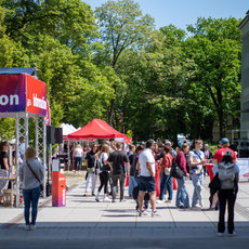 Menschenmenge auf einer Informationsveranstaltung im Freien mit rotem Zelt und mehreren roten Zelten im Hintergrund, bei der Besucher an Ständen und unter einem großen Infostand mit dem Schriftzug „Information“ stehen und sich unterhalten.