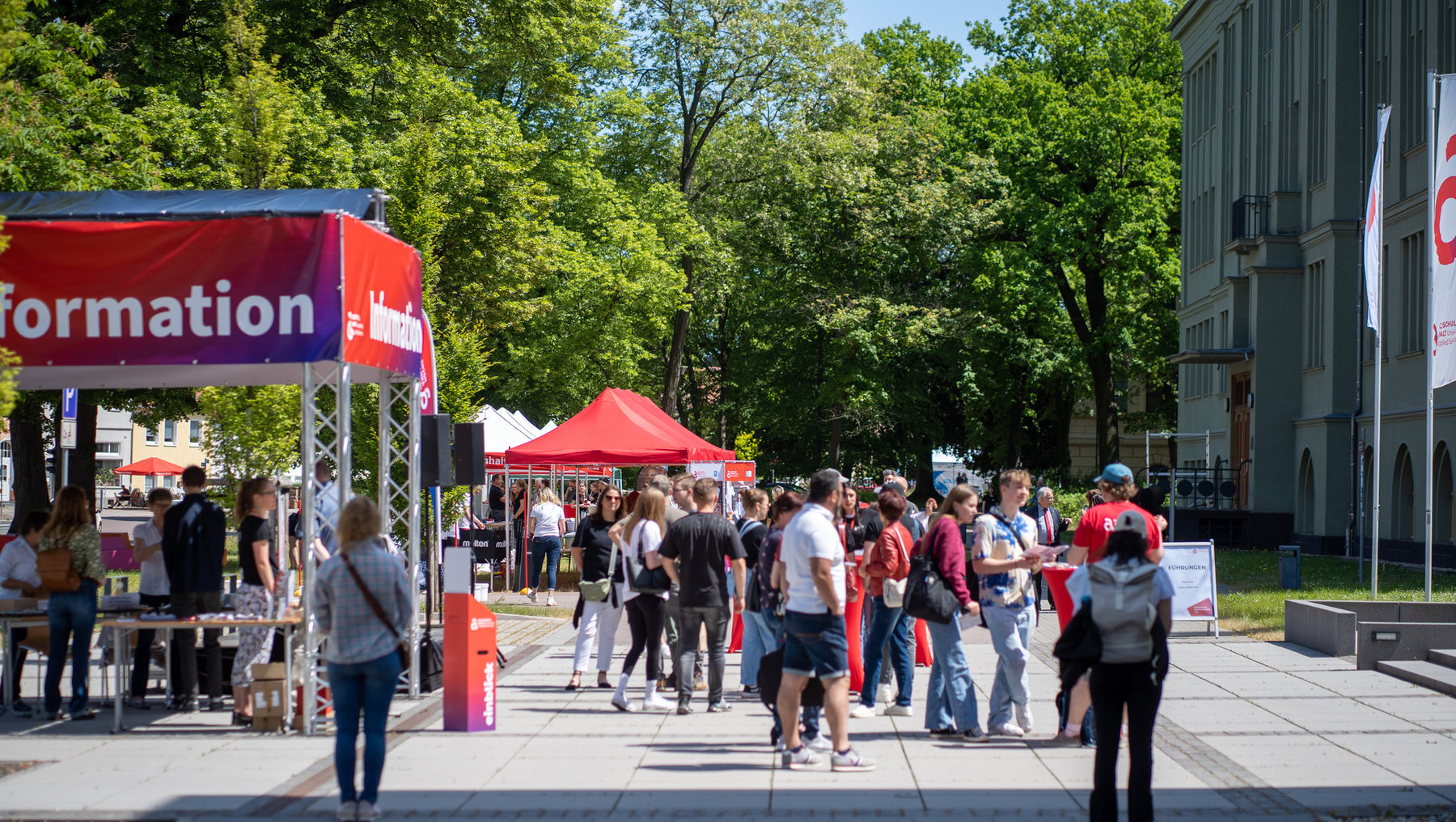 Menschenmenge auf einer Informationsveranstaltung im Freien mit rotem Zelt und mehreren roten Zelten im Hintergrund, bei der Besucher an Ständen und unter einem großen Infostand mit dem Schriftzug „Information“ stehen und sich unterhalten.