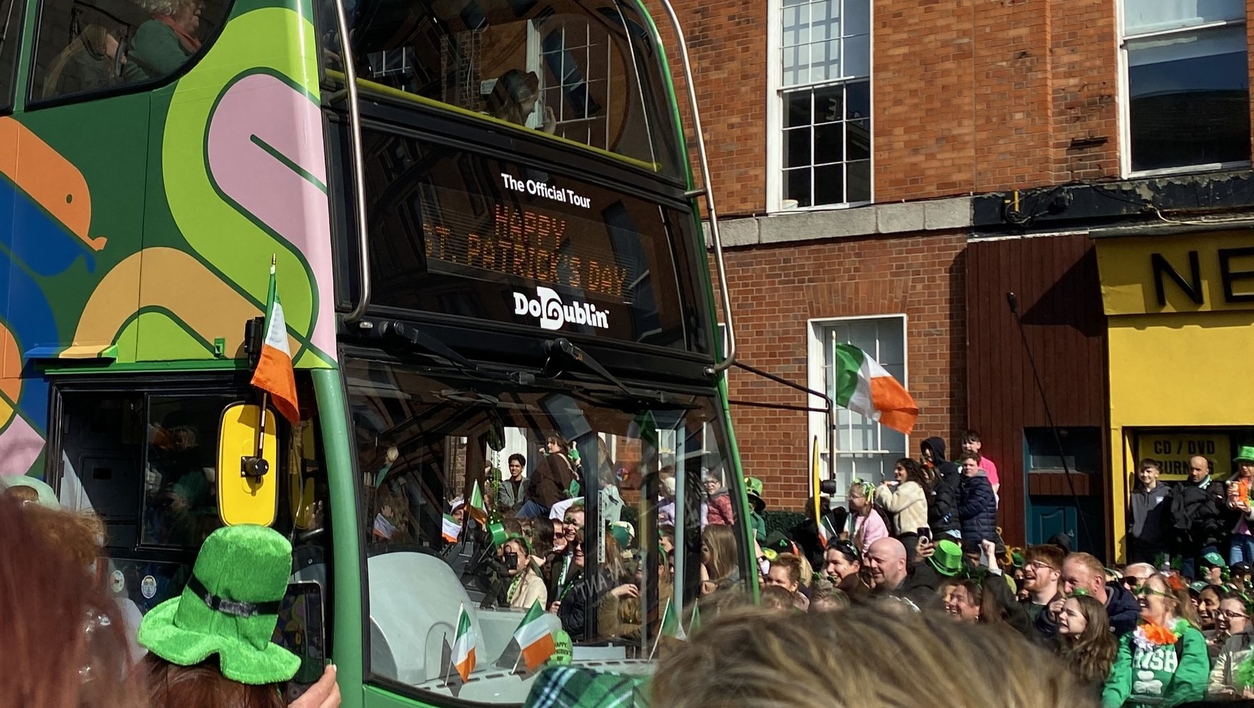 Bunter Doppeldeckerbus mit irischen Motiven wie Kleeblättern und grüner Bemalung fährt durch eine jubelnde Menschenmenge. Viele Zuschauer tragen grüne Kleidung und Accessoires, darunter Hüte mit Kleeblättern. Feierliche Stimmung auf einer Straße mit Gebäuden im Hintergrund.