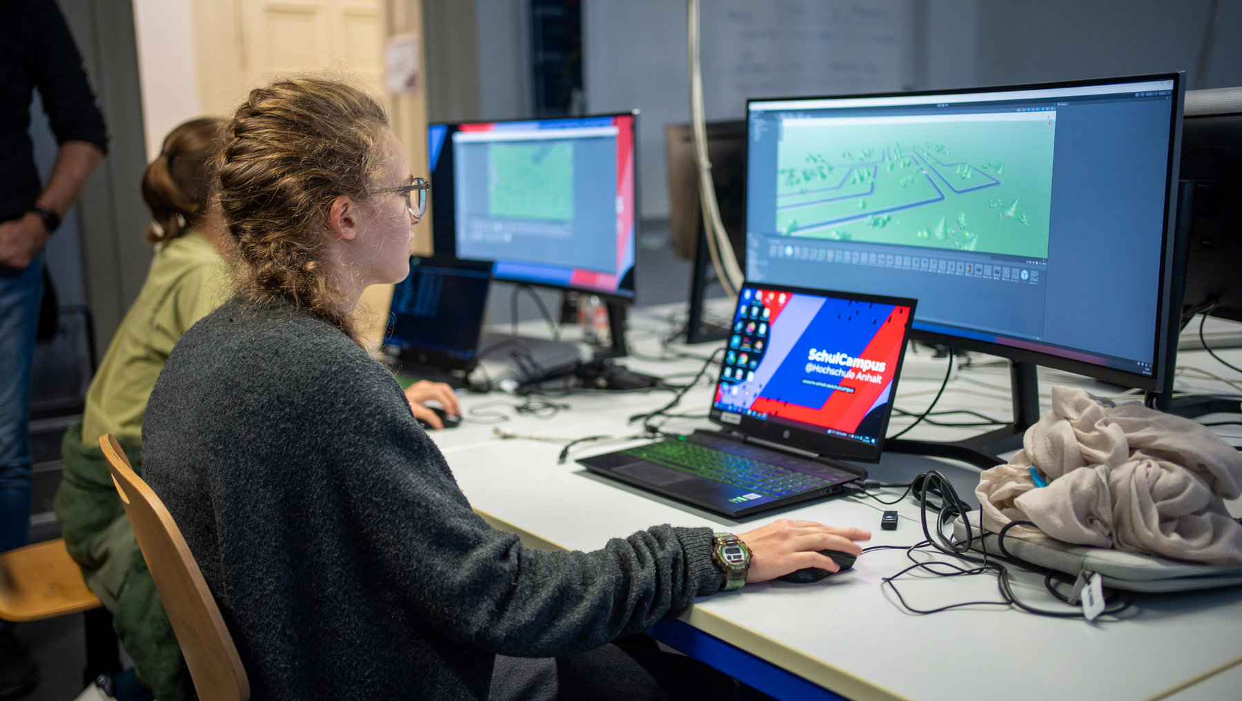Eine Person sitzt an einem Schreibtisch und arbeitet konzentriert an einem Laptop mit dem Aufdruck SchulCampus. Vor ihr stehen zwei Bildschirme, auf denen digitale Karten oder Pläne mit Gebäuden und Wegen zu sehen sind. Im Hintergrund arbeiten weitere Personen an ähnlichen Arbeitsplätzen.