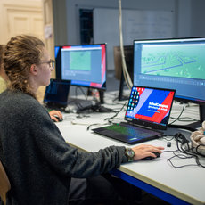Eine Person sitzt an einem Schreibtisch und arbeitet konzentriert an einem Laptop mit dem Aufdruck SchulCampus. Vor ihr stehen zwei Bildschirme, auf denen digitale Karten oder Pläne mit Gebäuden und Wegen zu sehen sind. Im Hintergrund arbeiten weitere Personen an ähnlichen Arbeitsplätzen.