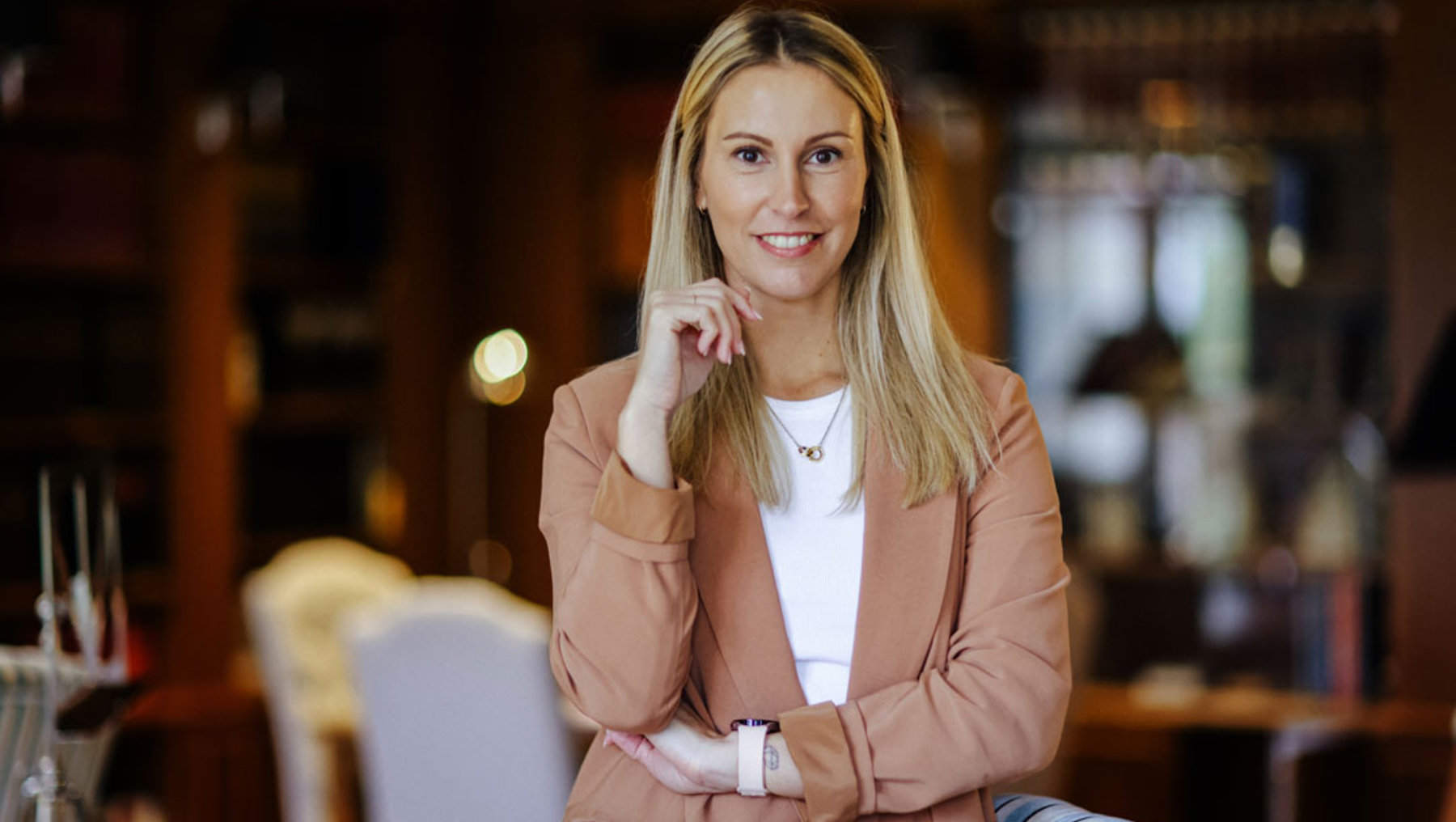 Eine lächelnde Frau mit hellblonden Haaren sitzt entspannt an einem Tisch in einem Restaurant oder Café, trägt eine beige Jacke über einem weißen Shirt und hält ein Handy in der Hand.