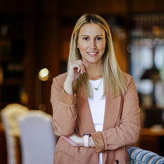 Eine lächelnde Frau mit hellblonden Haaren sitzt entspannt an einem Tisch in einem Restaurant oder Café, trägt eine beige Jacke über einem weißen Shirt und hält ein Handy in der Hand.