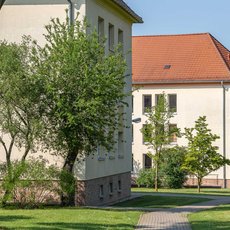 A large, two-story building with a red-tiled roof and white walls, surrounded by a well-maintained lawn with trees and a paved walkway. The building has multiple windows and appears to be a residential or institutional structure. The scene is bright and sunny.